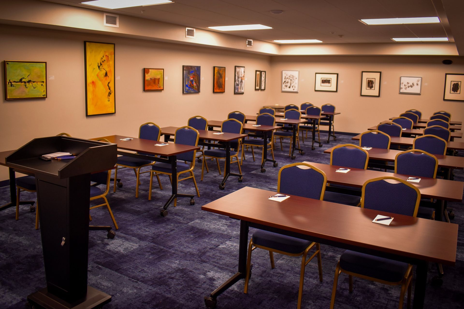 Empty conference room with rows of blue chairs, tables, podium, and framed artwork on beige walls