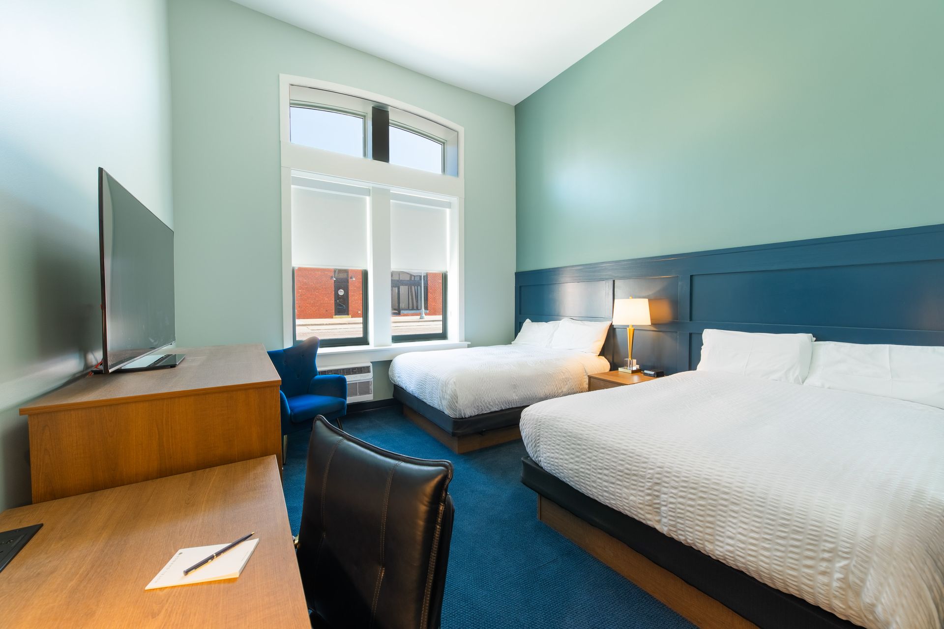 Modern hotel room with two beds, blue headboard, desk, TV, and a window overlooking buildings