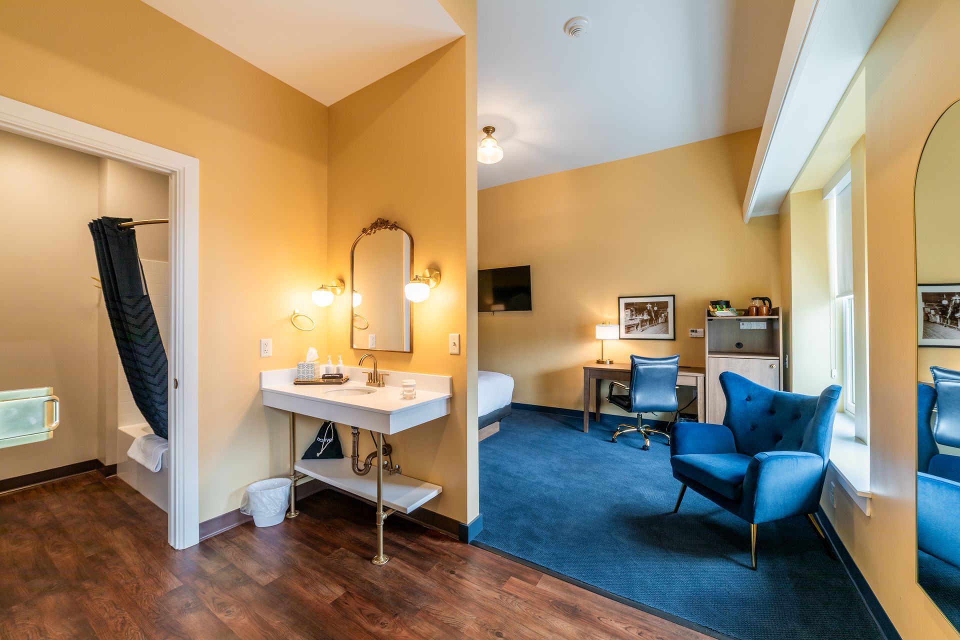 Yellow hotel room with desk, mirror, blue chair, sofa, and open doorway to bathroom