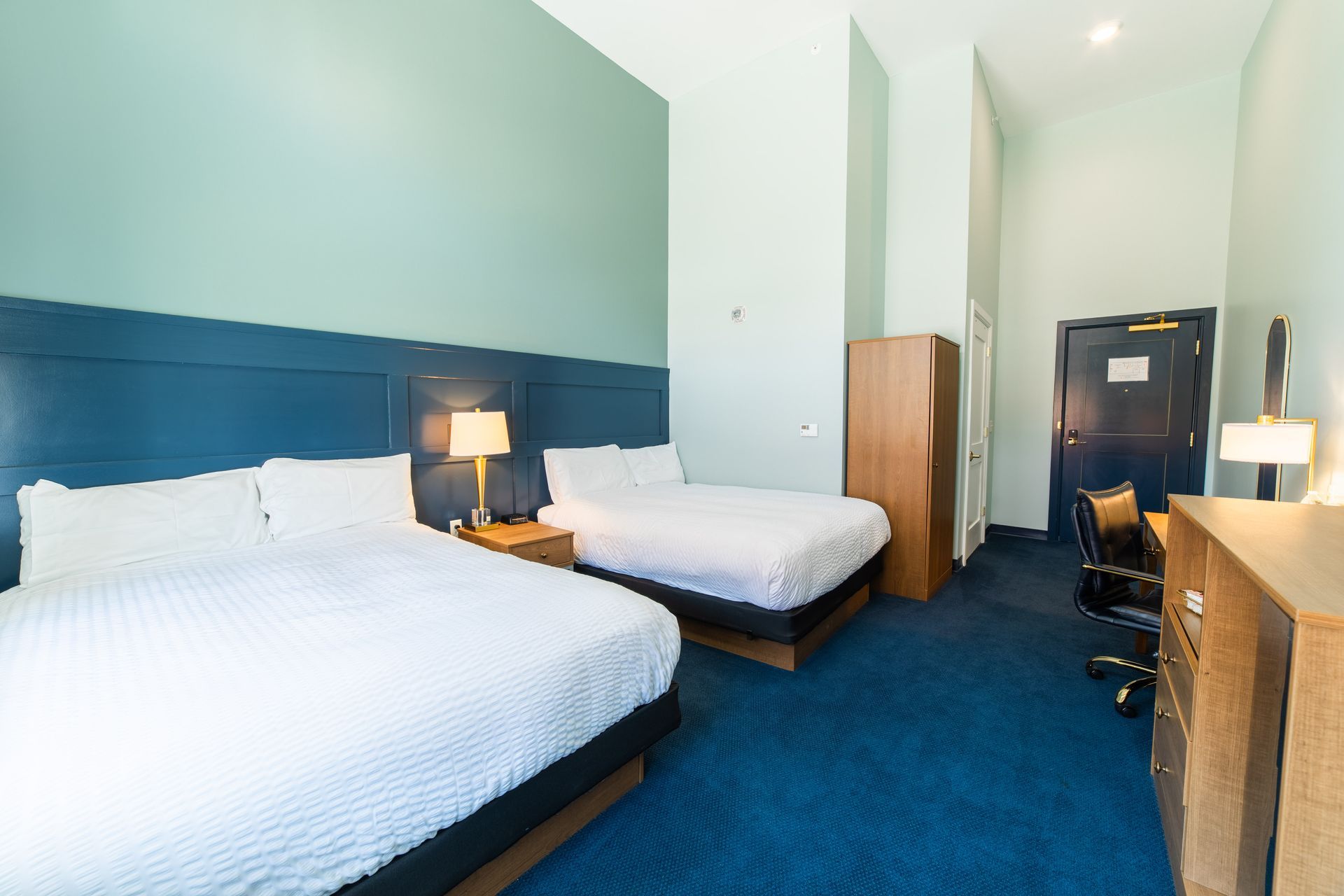 Hotel room with two white beds, blue carpet, desk, and lamp in a bright, modern space