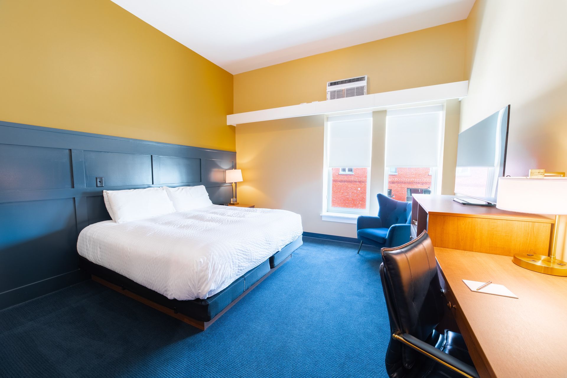 Modern hotel room with a king bed, blue carpet, desk, chair, and balcony doors.