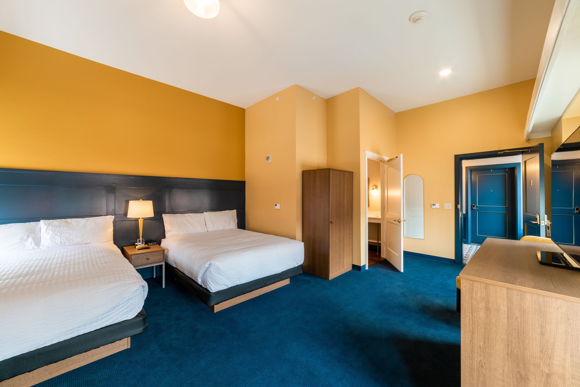 Hotel room with two beds, blue carpet, yellow walls, and a desk near the window.