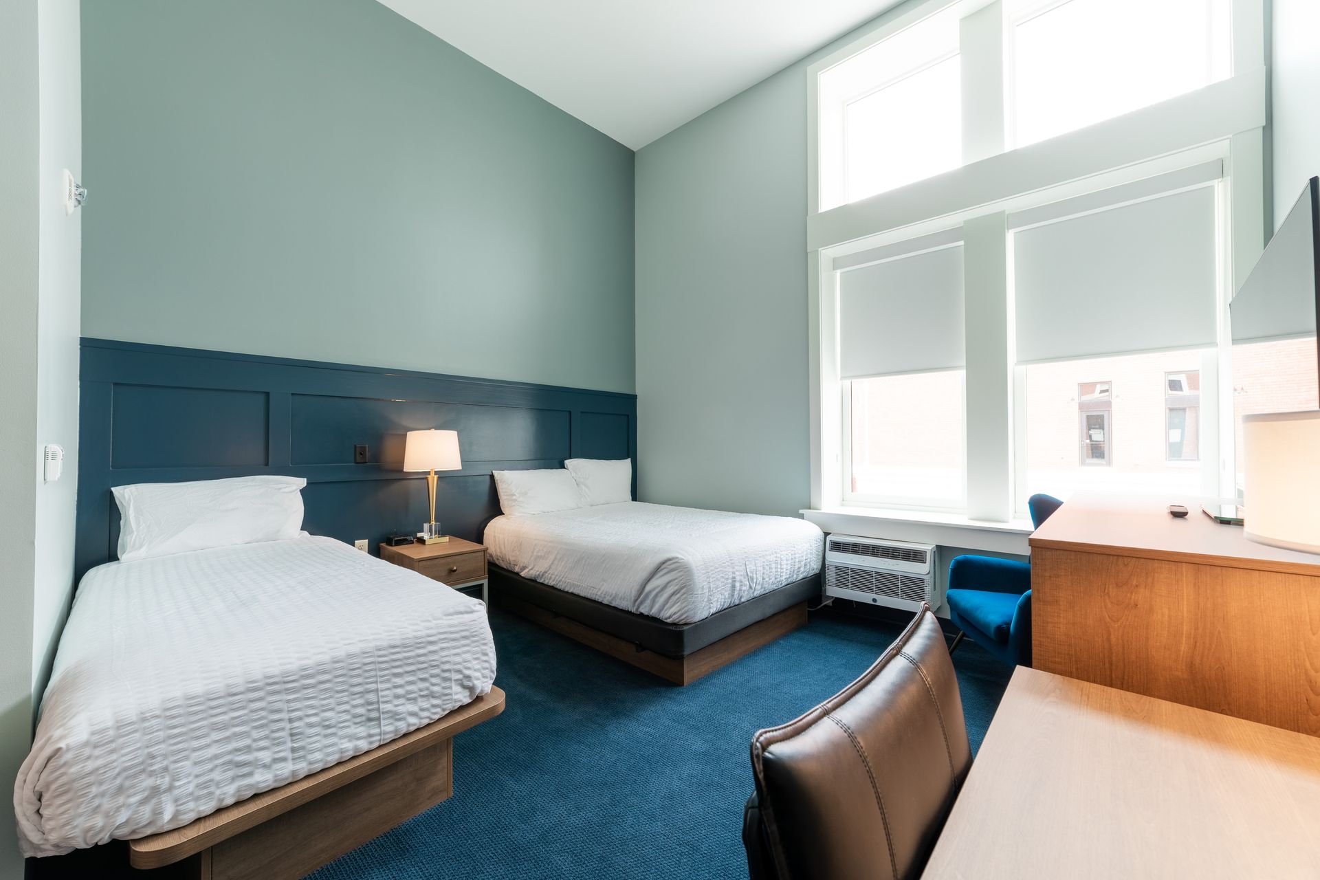 Dorm room with two beds, blue carpet, desk, and large windows with blinds