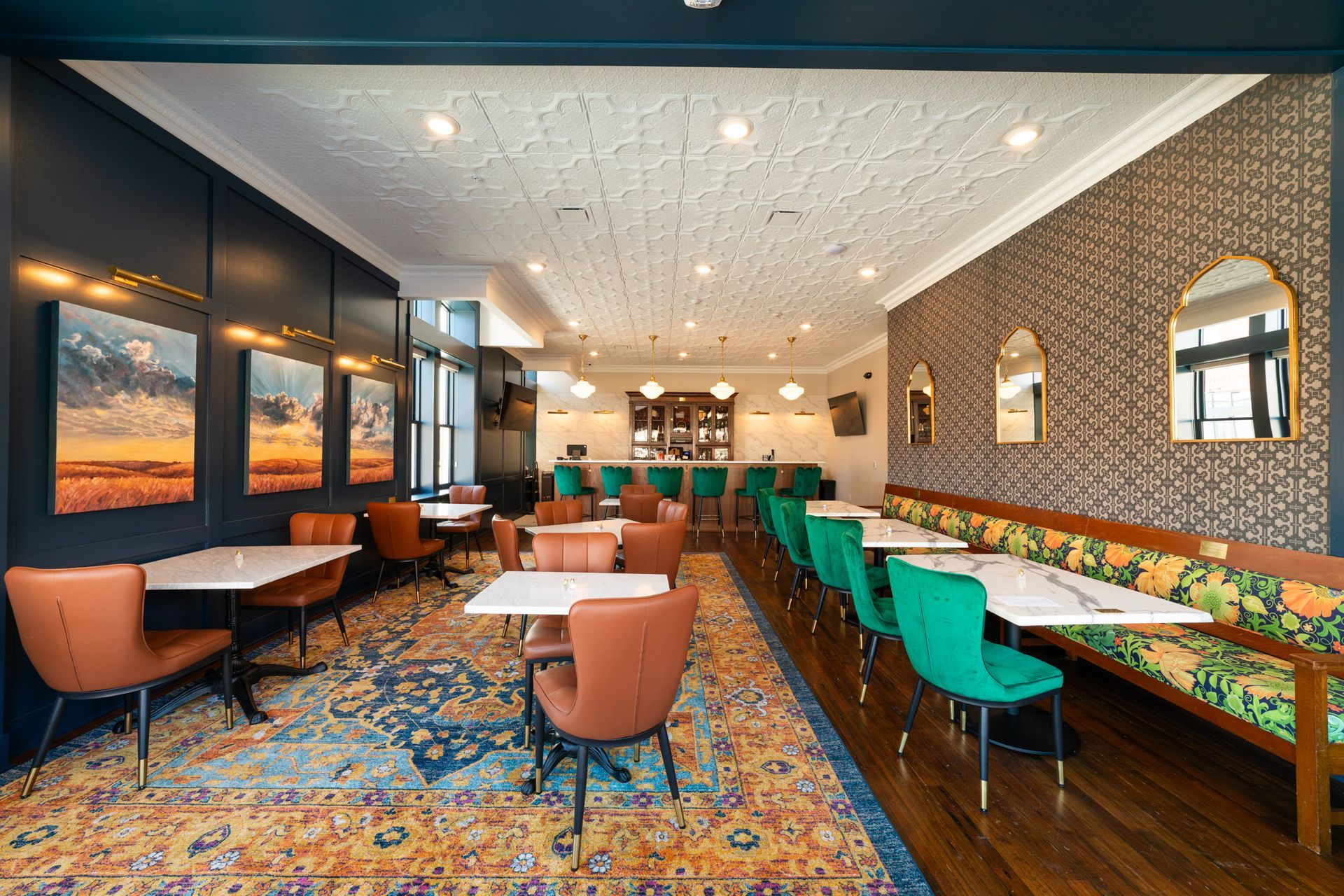 Restaurant interior with tables, chairs, bar, patterned ceiling, and artwork. Teal, orange, and gold colors.