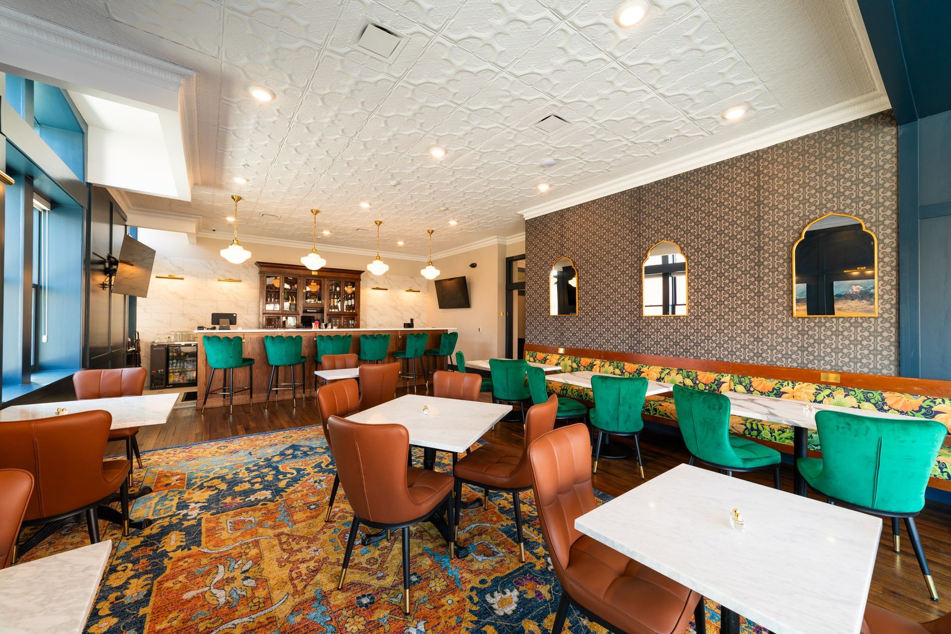 Restaurant interior with a bar, tables, and eclectic seating on a patterned rug.