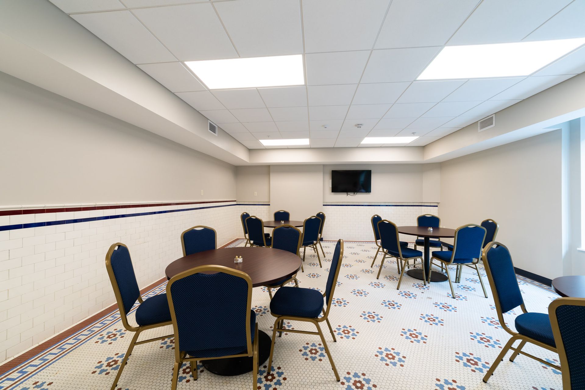 An empty conference room with round tables, blue chairs, a patterned floor, and a television mounted on the wall.