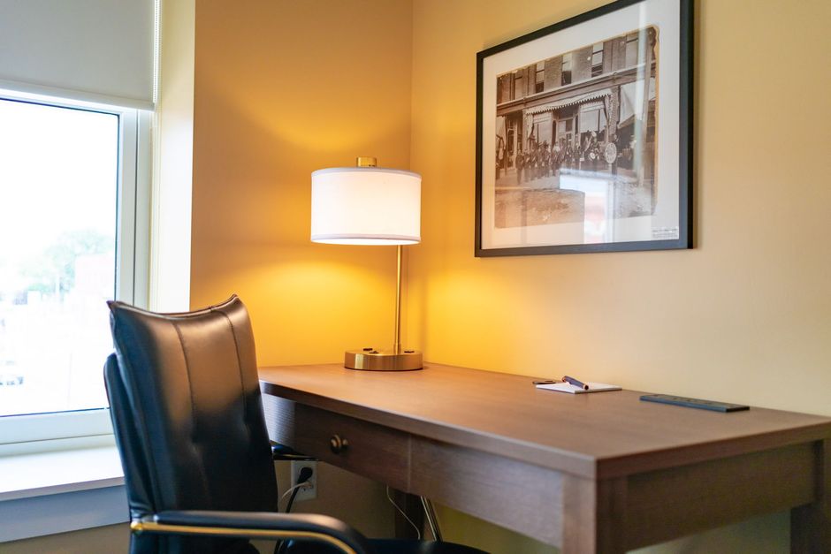 Desk with lamp, chair, and framed artwork near a window in a room with beige walls.