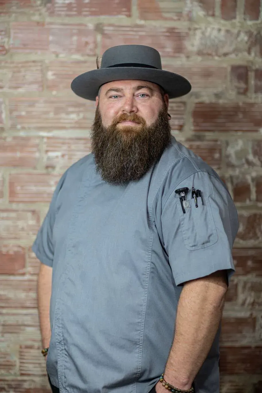 Bearded person in a gray shirt and black hat standing against a brick wall.