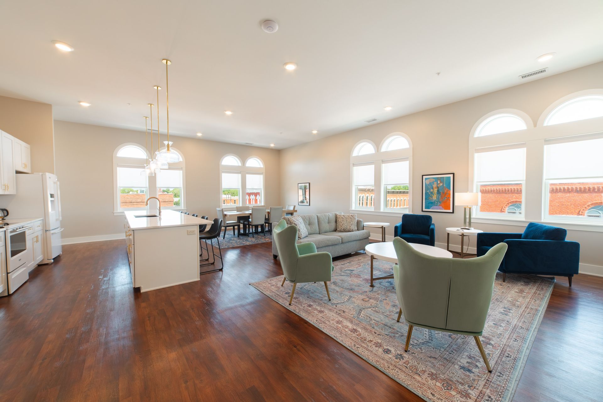 Bright open living room with hardwood floors, large windows, blue and green chairs, and a kitchen island.