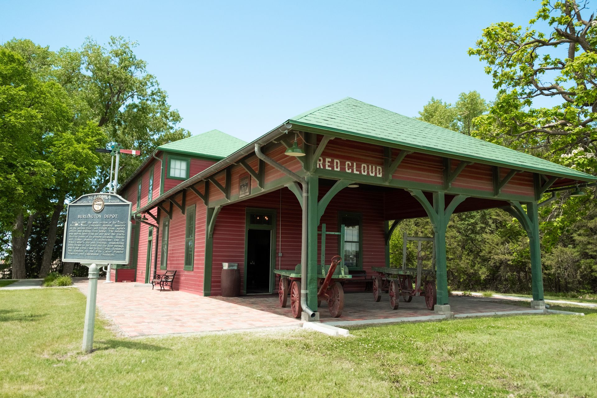 Red brick historic train depot with green roof and porch, surrounded by trees and grass