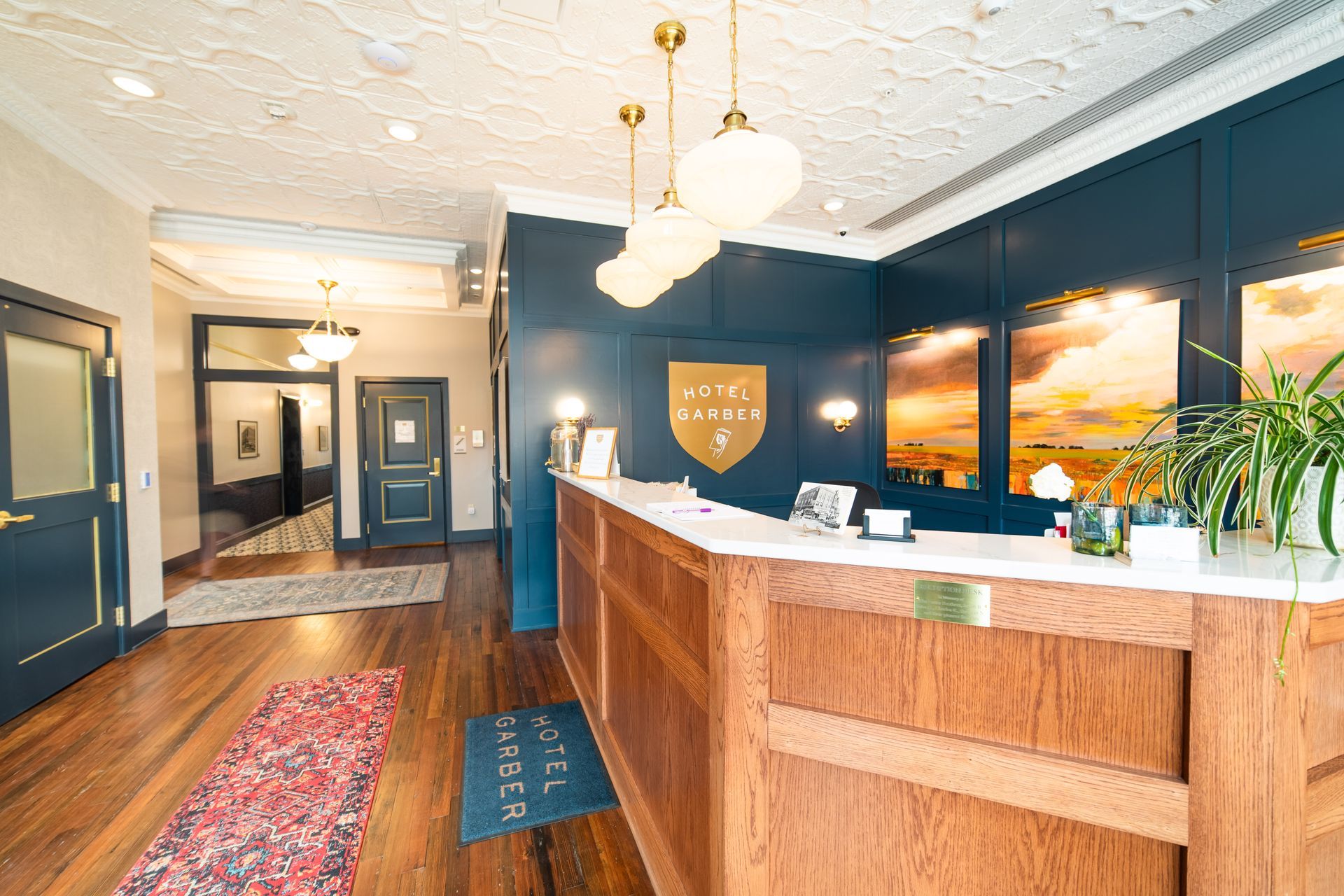 Hotel reception lobby with wood desk, dark blue walls, patterned rug, and hanging lights