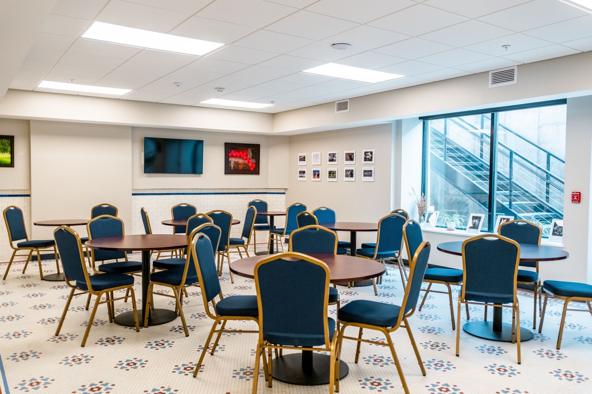 Empty conference room with blue chairs, round tables, wall screens, and a large window overlooking stairs