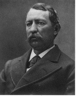 Man in dark suit, with mustache, looking directly at the viewer.