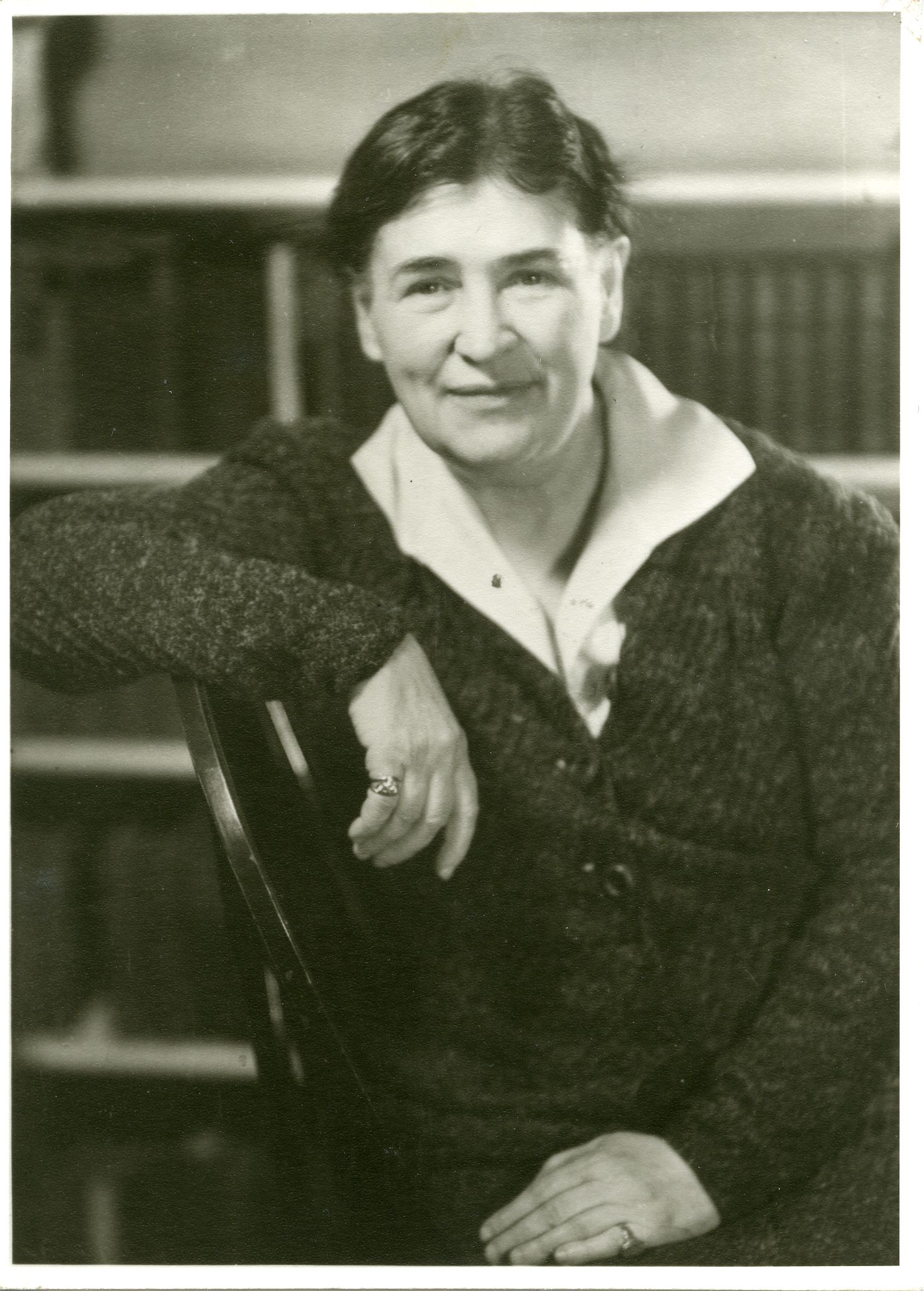 Woman seated, leaning on chair, in front of a bookshelf. She is wearing a dark sweater and looking at the camera.