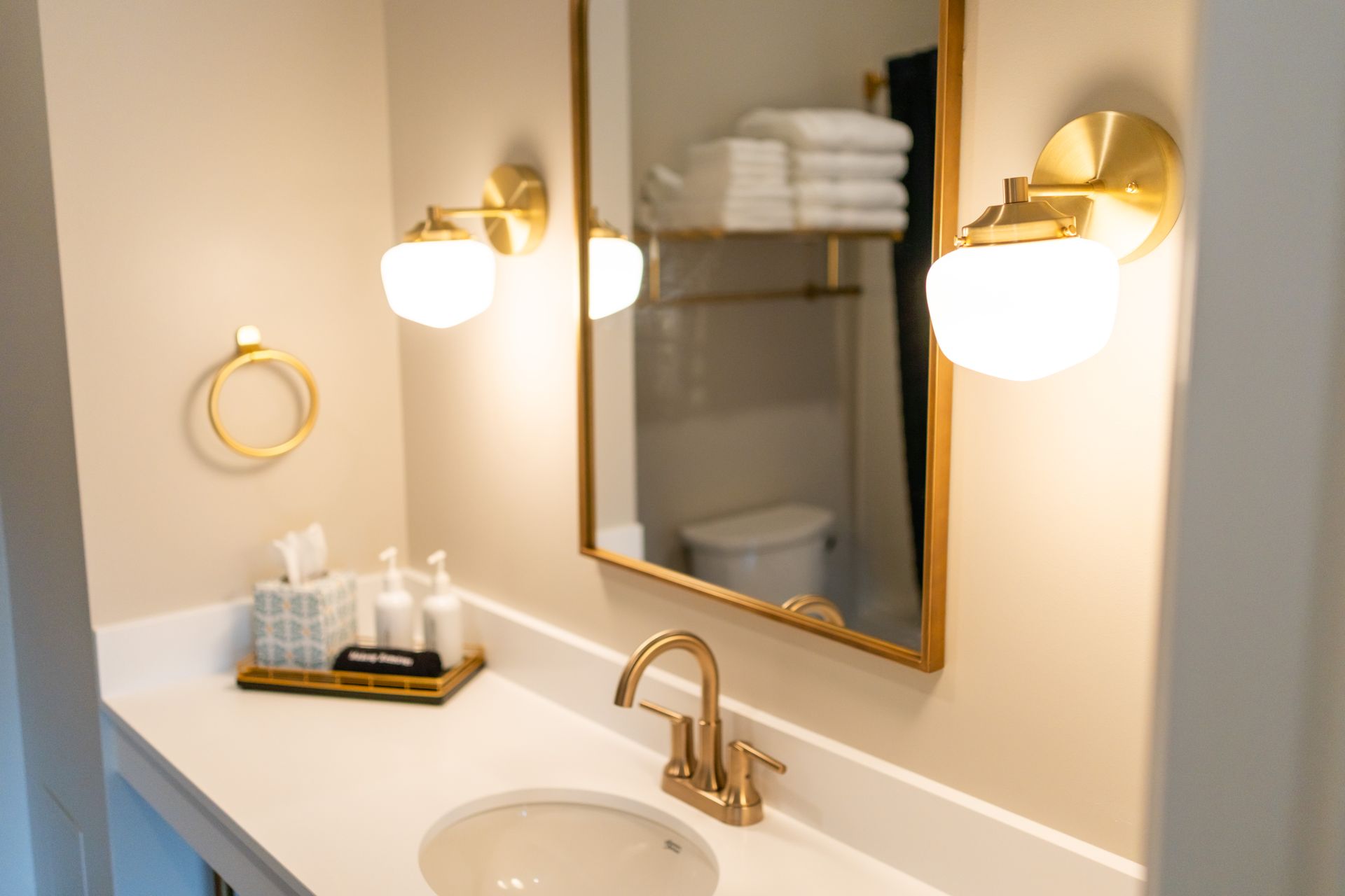 Modern bathroom vanity with gold fixtures, mirror, wall sconces, and neatly stacked towels