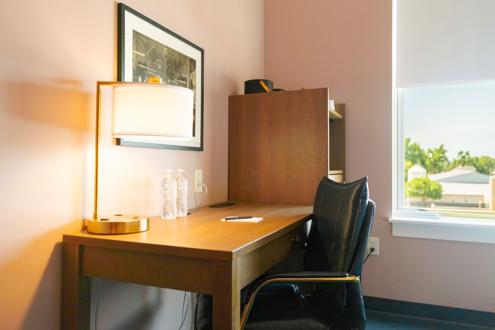 Small office desk with lamp and chair beside a window, with a wooden cabinet and framed art.