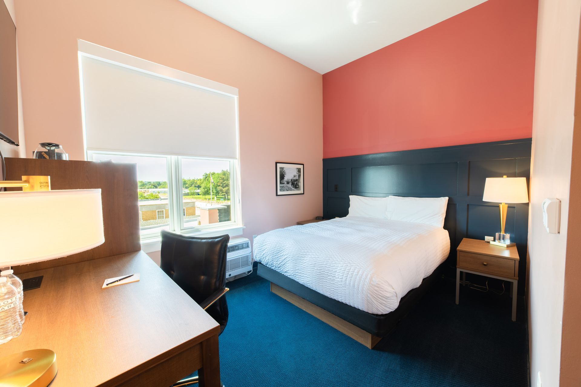 Hotel room with a bed, desk, and two lamps, featuring peach walls and blue carpet.