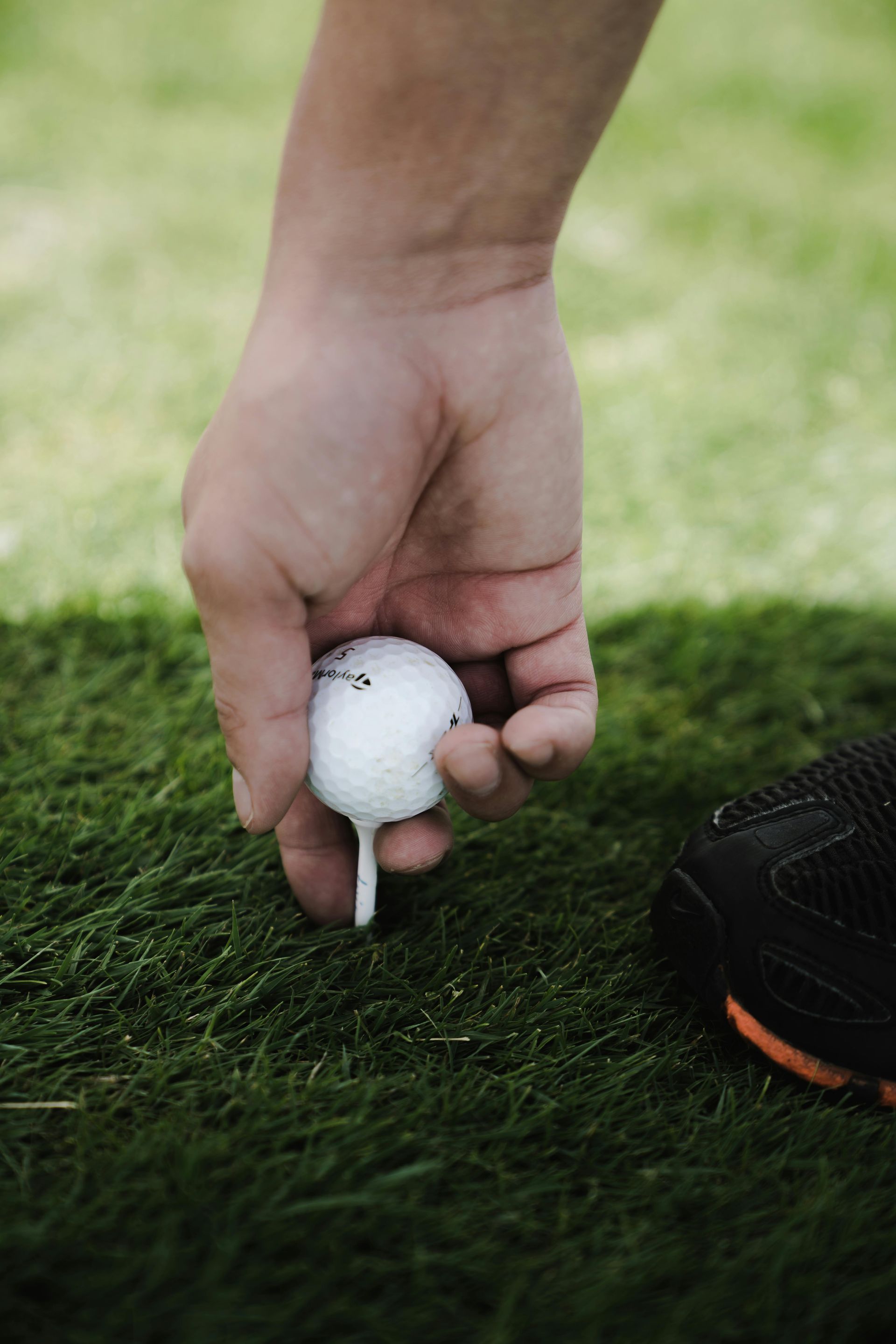A hand places a white golf ball on a tee in the green grass, with a golf shoe visible in the corner.