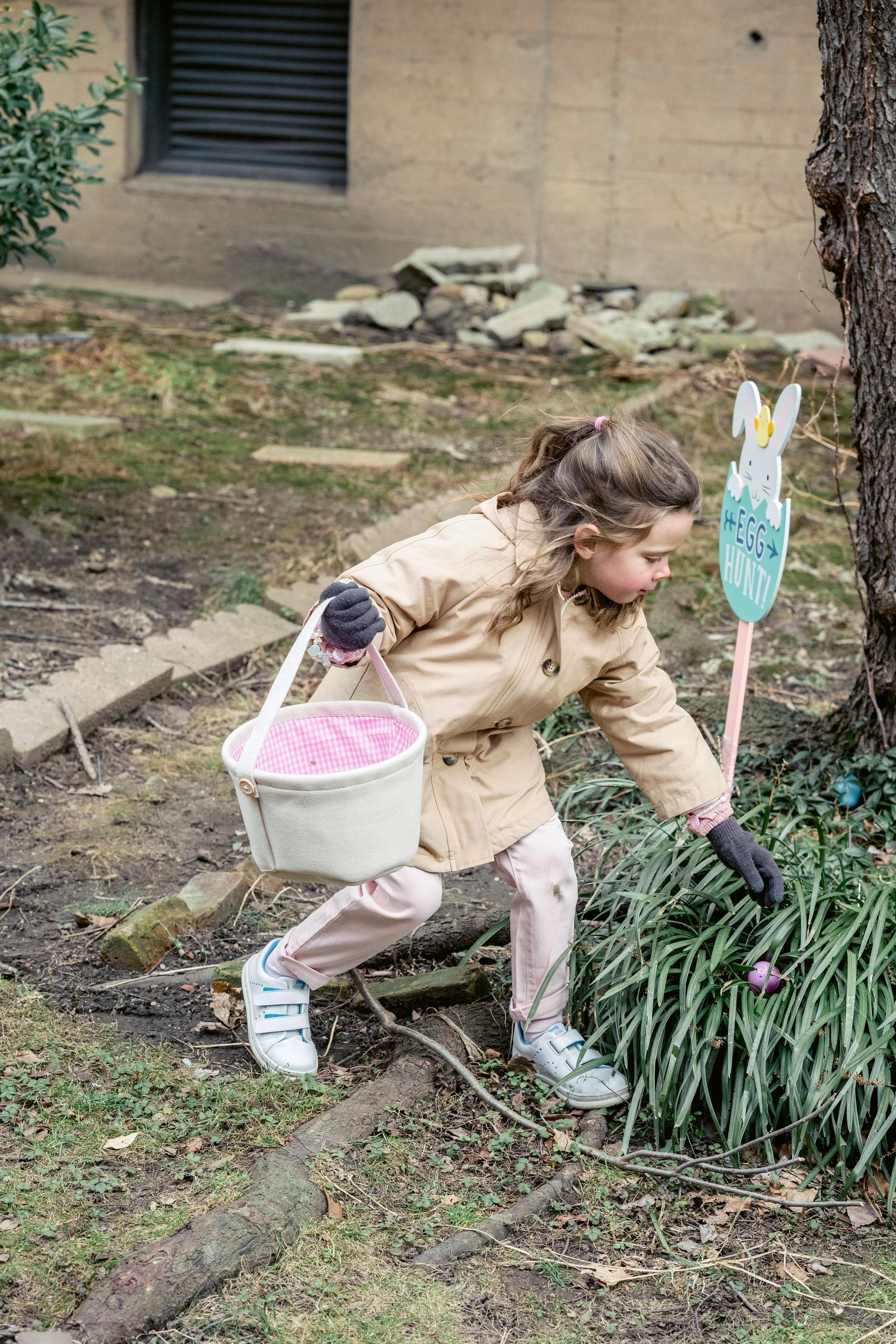 Girl in beige jacket reaches for an egg while holding a pink Easter basket near a tree in a yard.