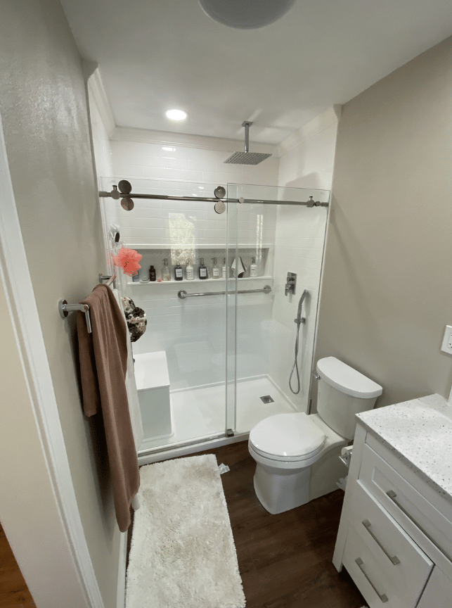 Small bathroom with glass shower, toilet, vanity, towel rack, and white rug.