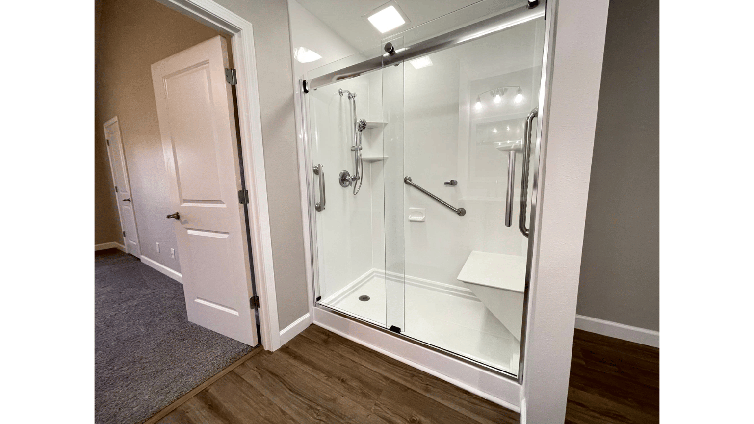 Modern accessible shower with glass doors in a hallway beside a beige door and wood flooring