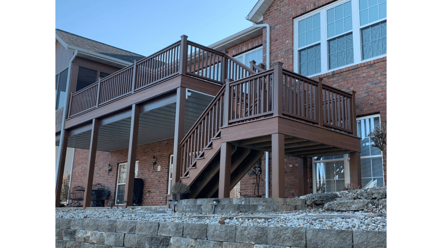 Brown wooden deck and stairs attached to a brick house, viewed from a stone retaining wall.
