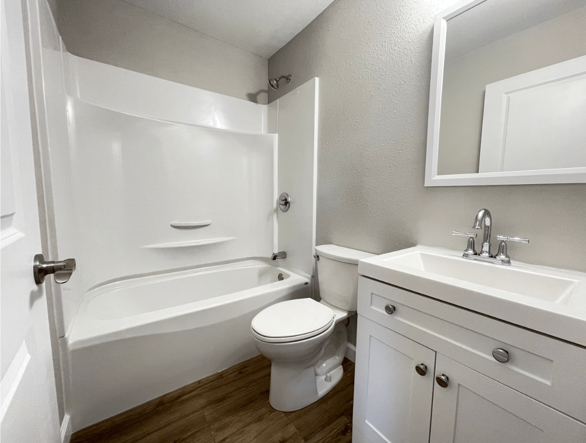 White bathroom with tub-shower, toilet, and vanity sink with mirror; wood-look floor.