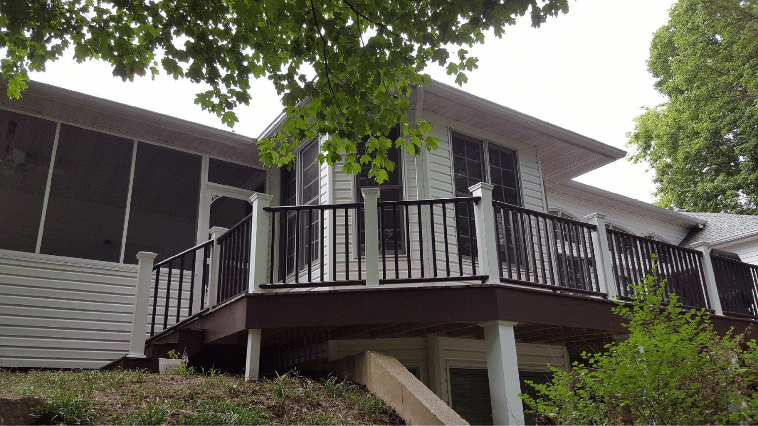 House with a raised wooden deck and black railing, surrounded by trees and greenery