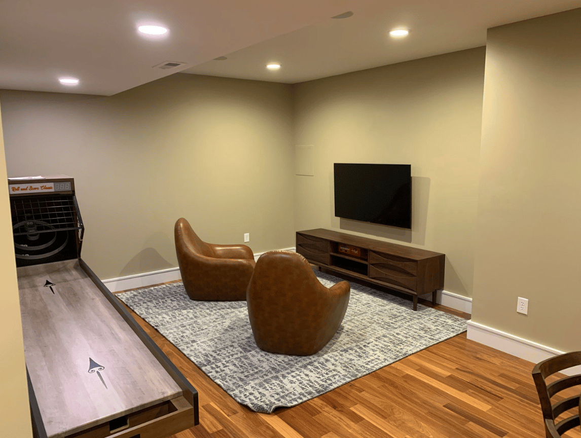 Basement lounge with TV, two brown chairs, rug, hardwood floor, and a long bench or table on the left.