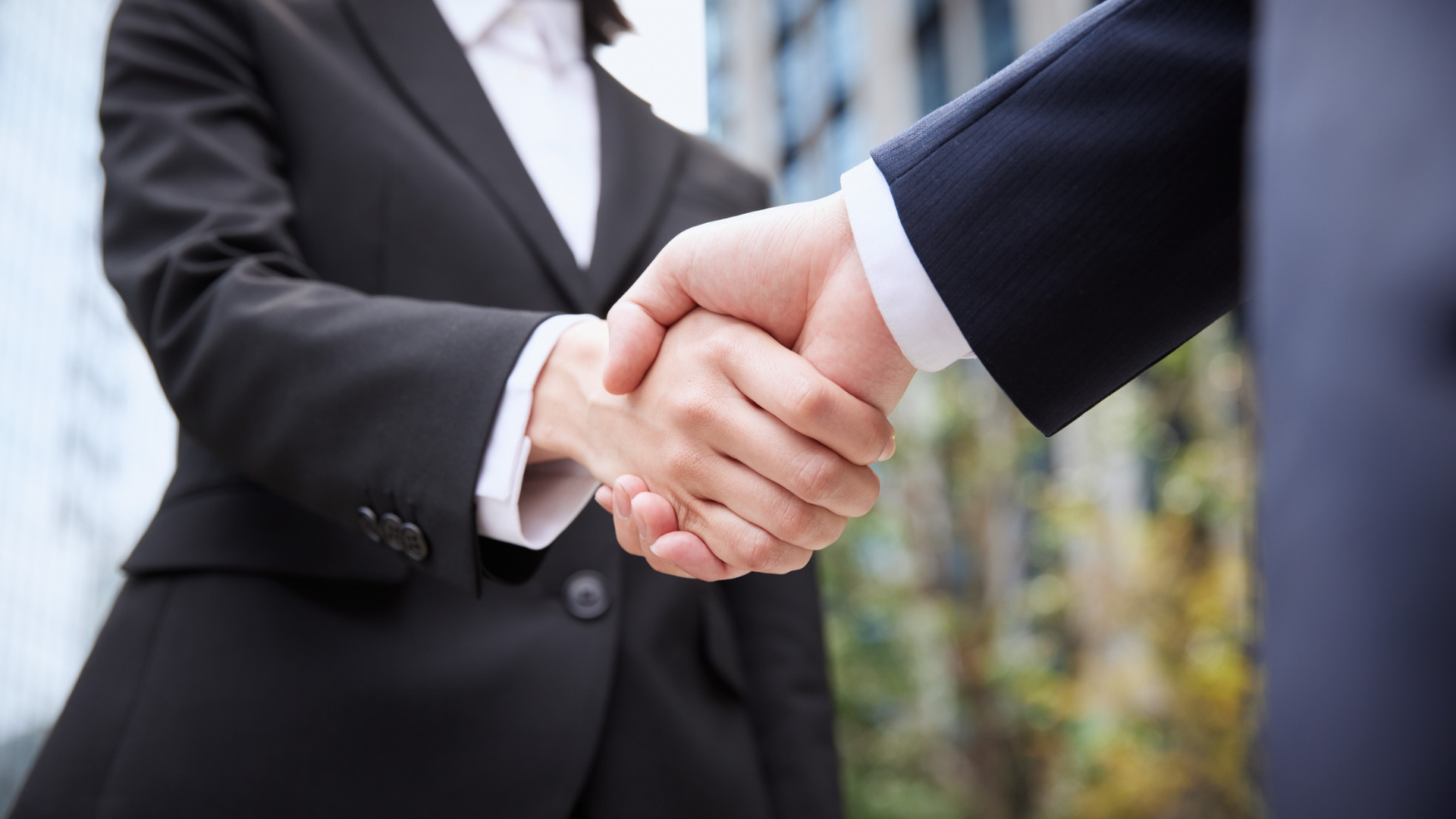 Two businesspeople shaking hands outdoors, with office buildings blurred in the background.