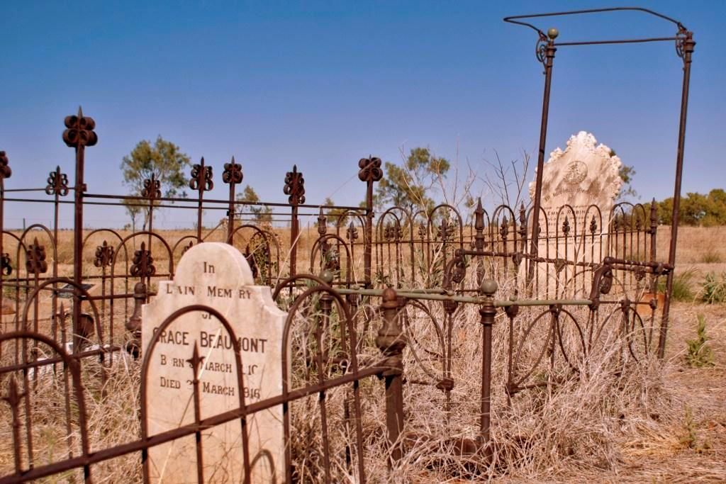 Old desert cemetery with wrought-iron fence, weathered headstones, and an arched mausoleum in the background