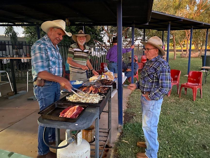Cowboys cooking at an outdoor grill under a pavilion, talking with a man in plaid