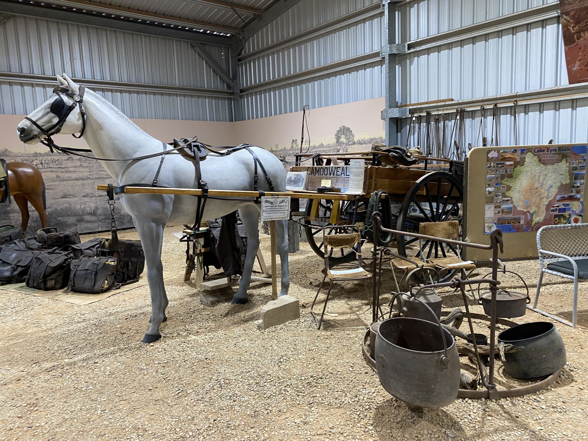 White horse harnessed to a wagon in a barn, with antique tools and equipment nearby.