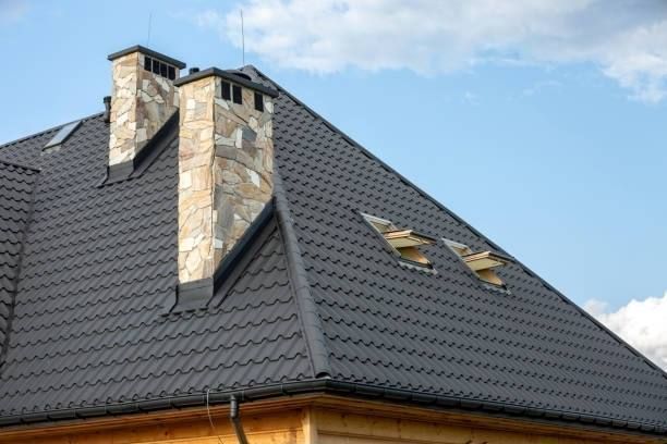 Dark gray metal roof with stone chimneys and skylights against a blue sky.