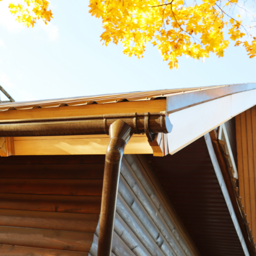 Brown rain gutter on a house with yellow leaves in the background under a blue sky.