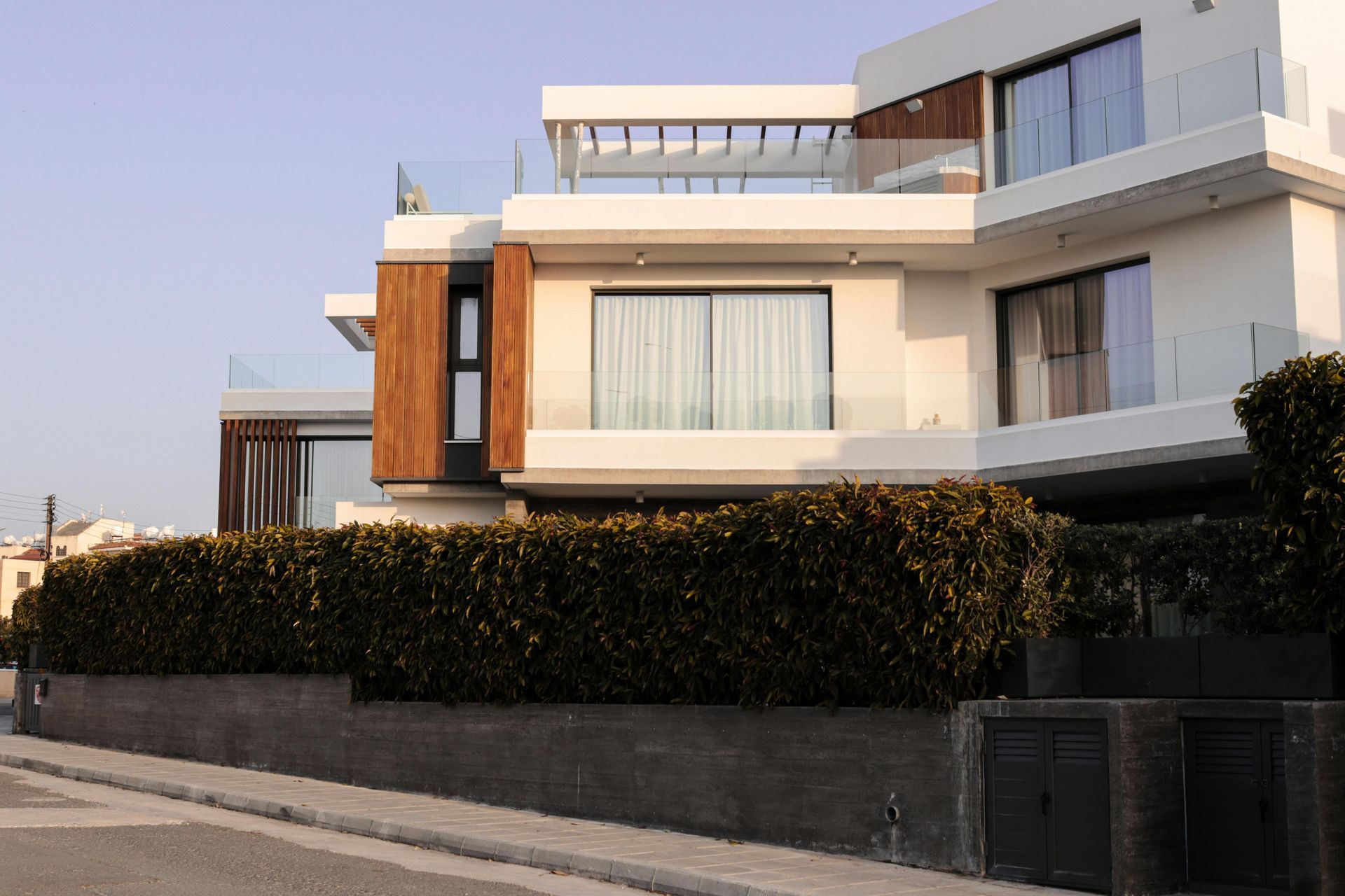 Modern multi-story house with wood accents, large windows, and a hedge along the street.