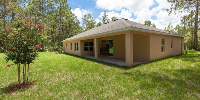 Single-story beige building with a green roof in a grassy yard, surrounded by trees and blue sky