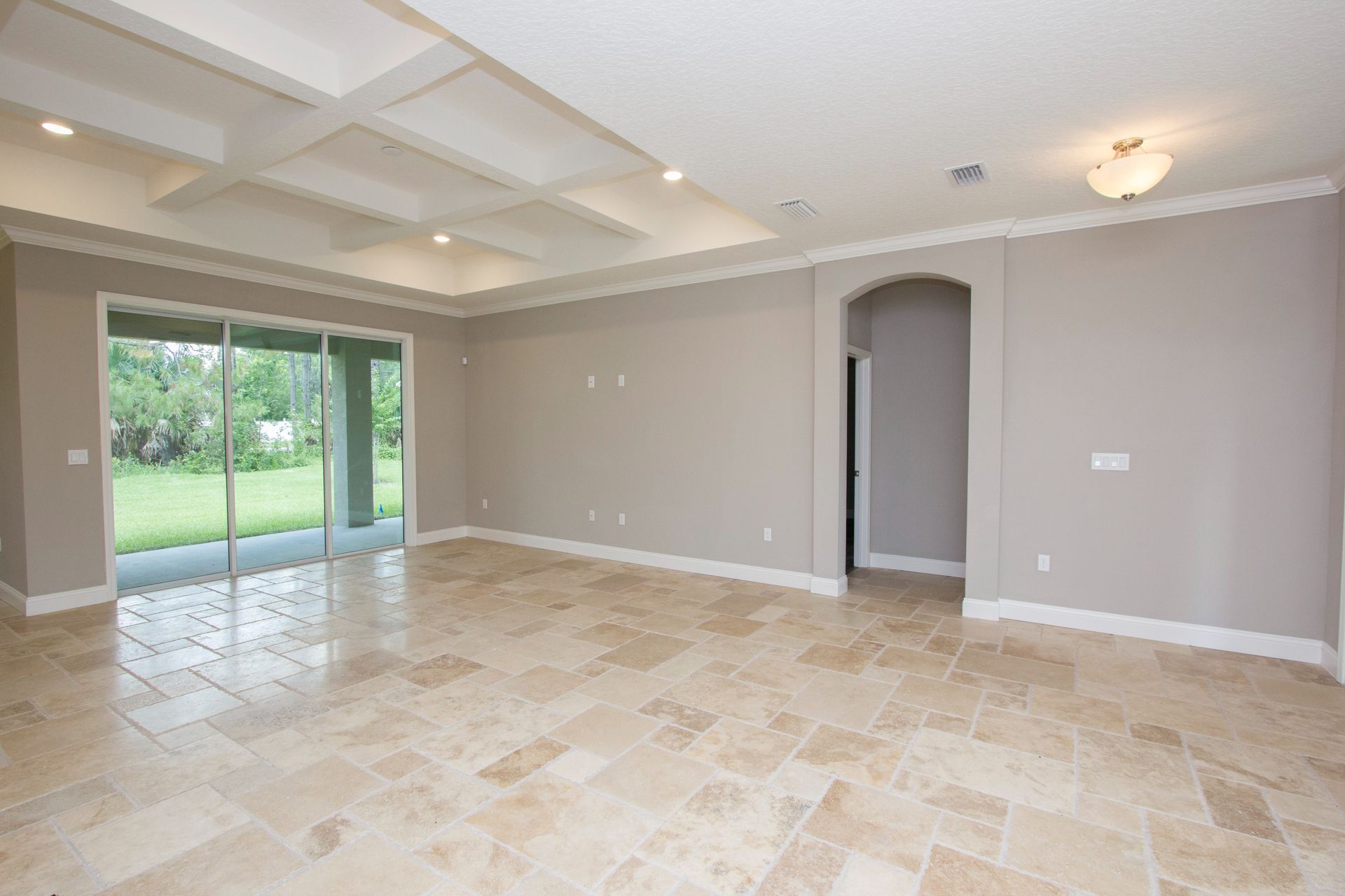 Empty beige living room with tiled floor, tray ceiling, arched doorway, and sliding glass doors to a green yard
