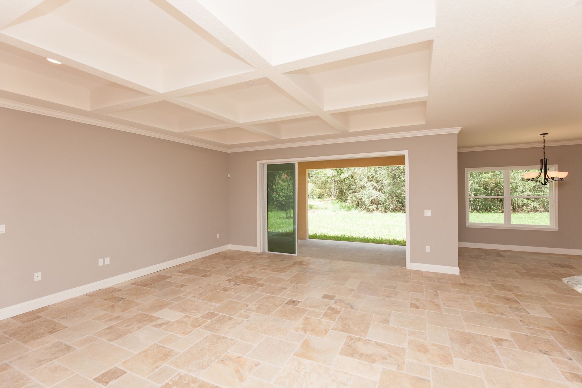 Empty beige living room with tiled floor, coffered ceiling, and sliding glass doors to a green yard