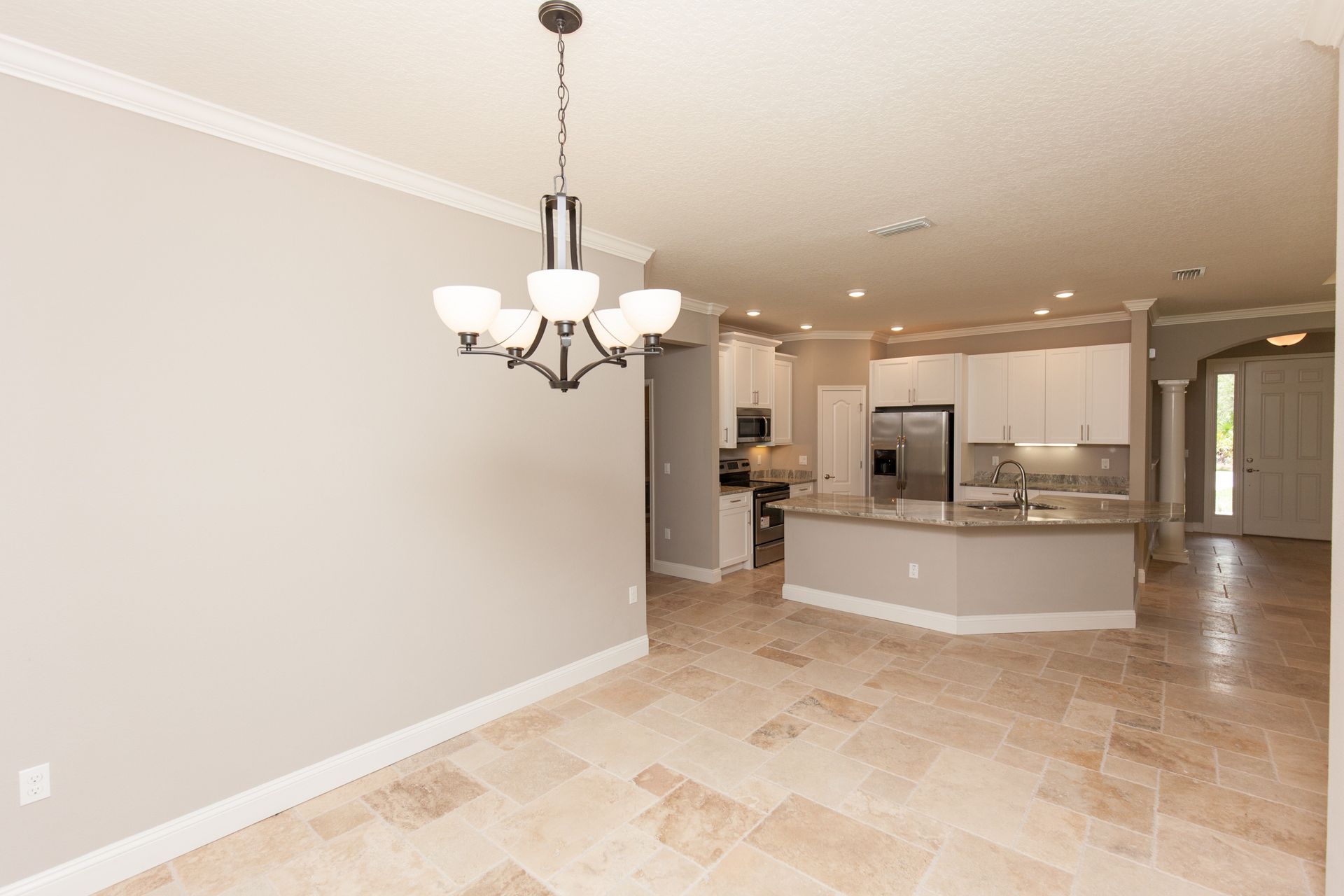 Empty beige dining and living area with tiled floor, chandelier, and open kitchen in the background