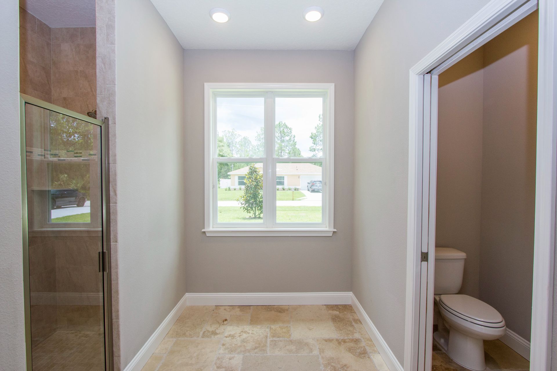 Bright bathroom with glass shower, window, beige tile floor, and toilet in a side alcove