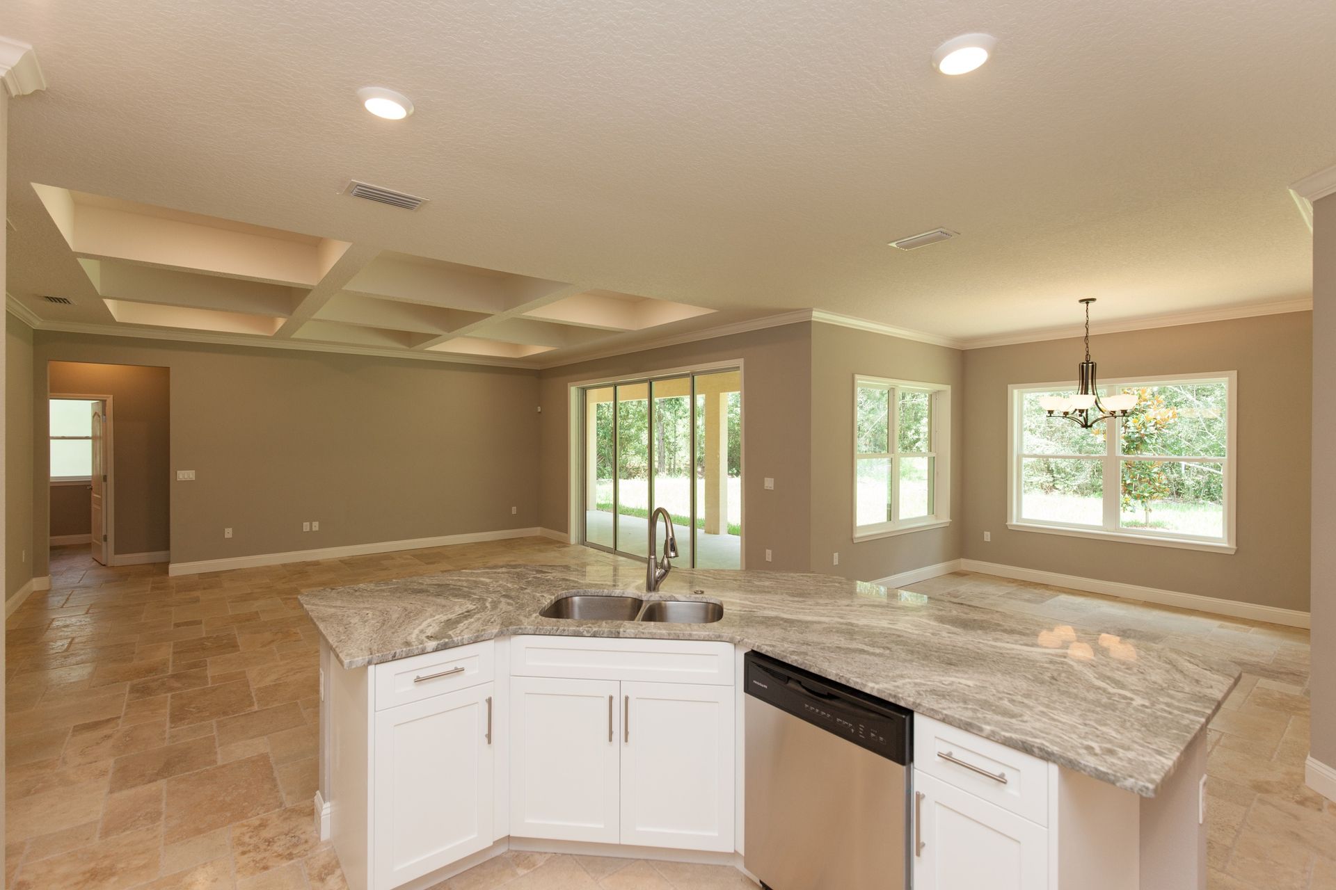 Spacious empty living room with beige tile floors, white cabinets, and large windows opening to greenery