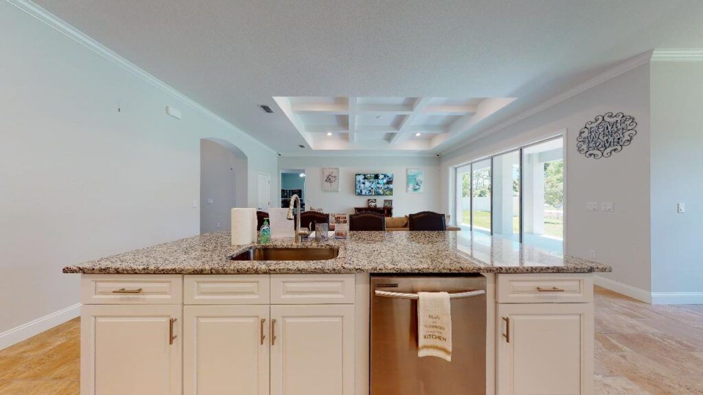 Bright open-plan kitchen with granite island, cream cabinets, and view into a sunlit living room and patio.