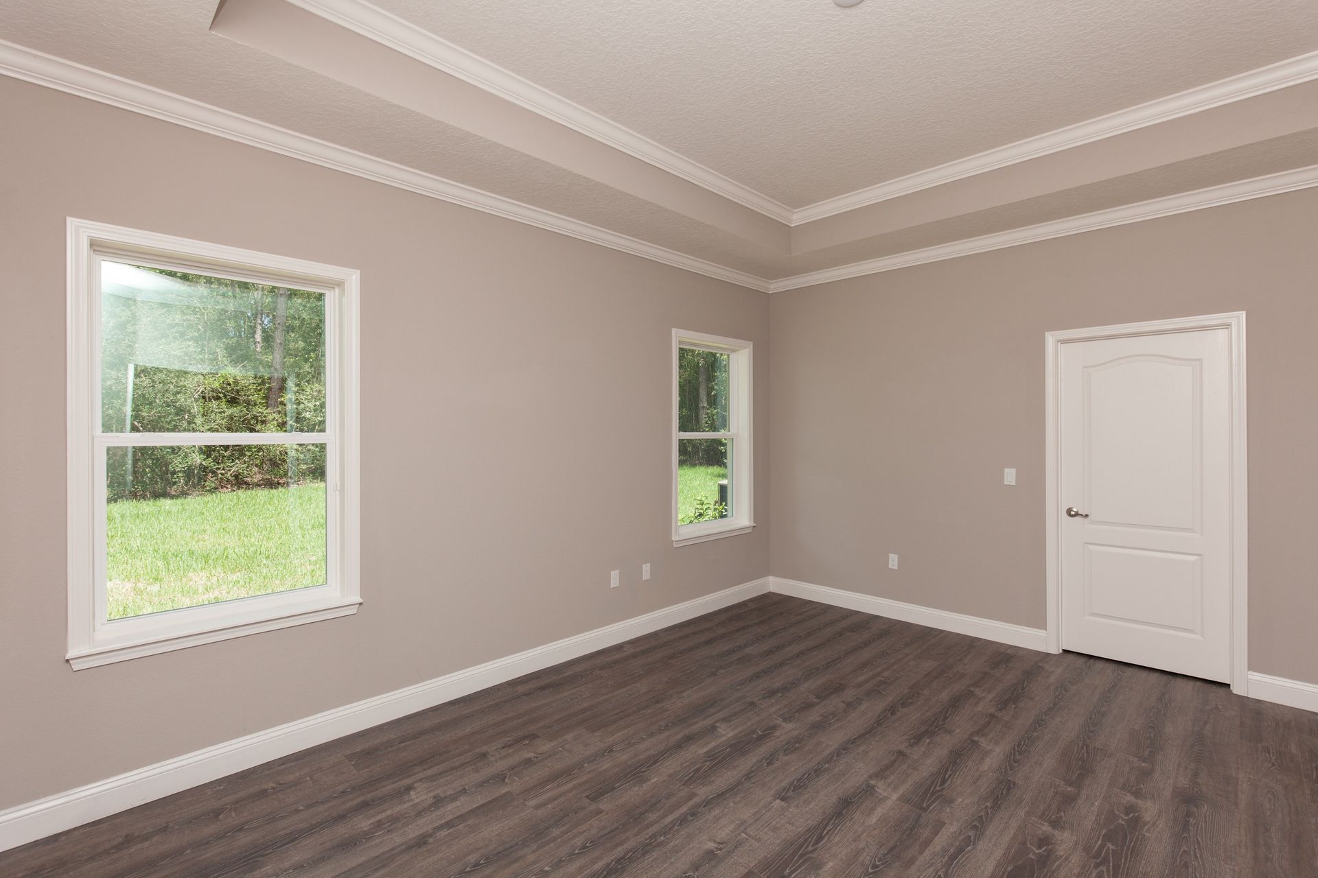Empty beige room with gray wood flooring, two windows, crown molding, and a white door