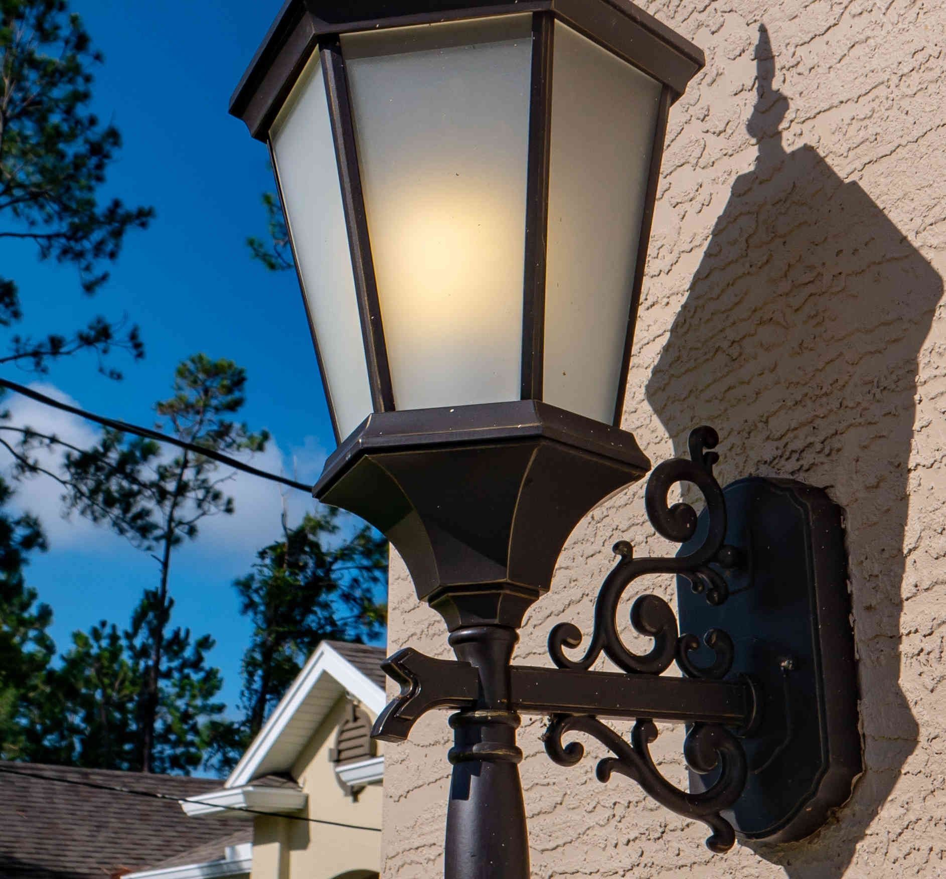 Black outdoor wall lantern on a stucco house at dusk with blue sky and trees in the background