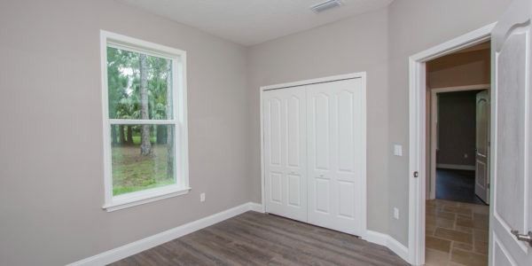 Empty light-gray bedroom with wood floor, window, double closet doors, and open doorway to a hall