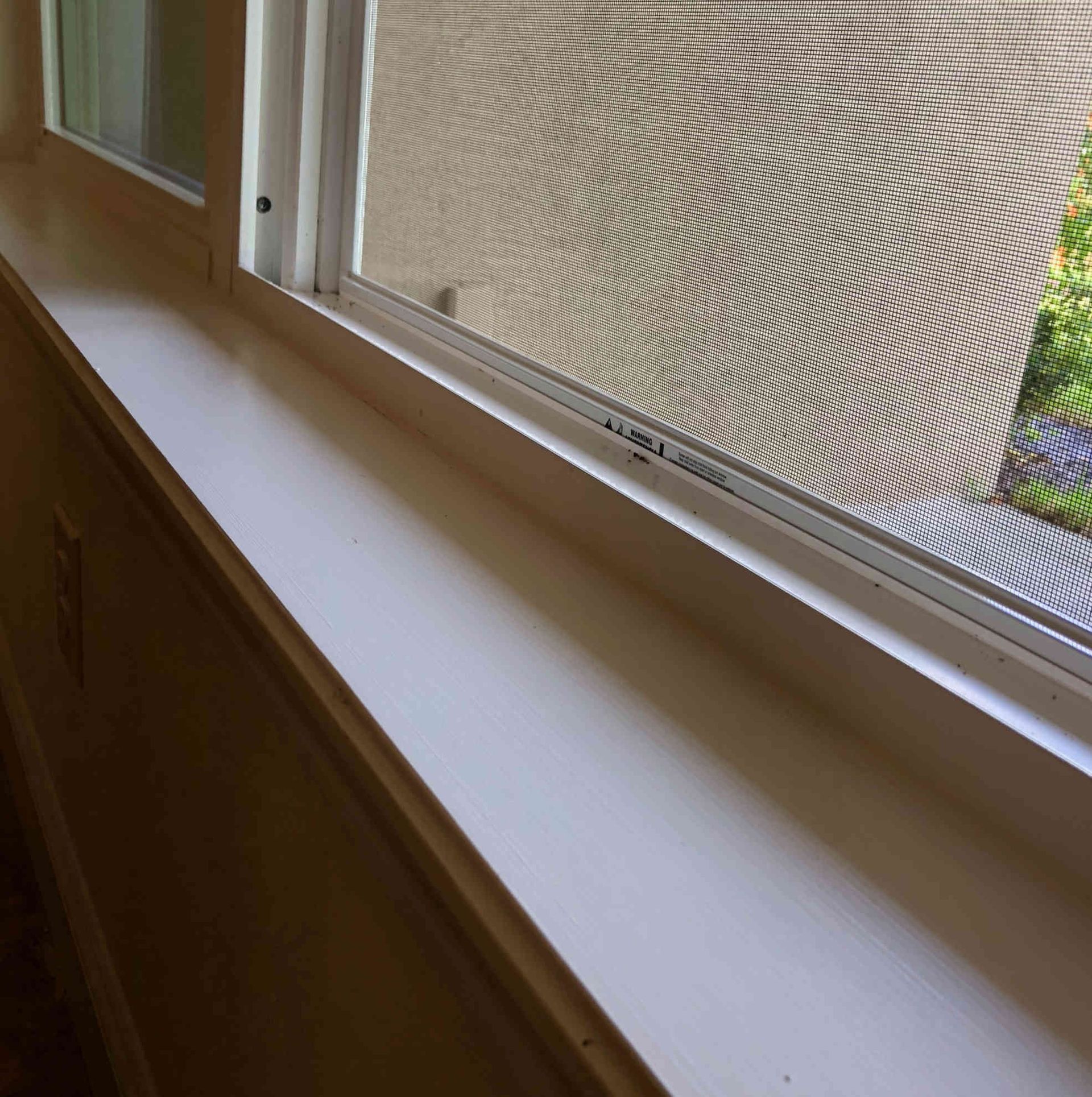 Window with a white sill and partially open blind, showing a view of greenery outside