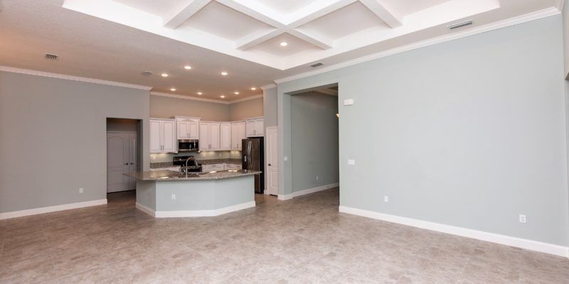 Empty modern living room with beige carpet, white walls, and a kitchen island in the background