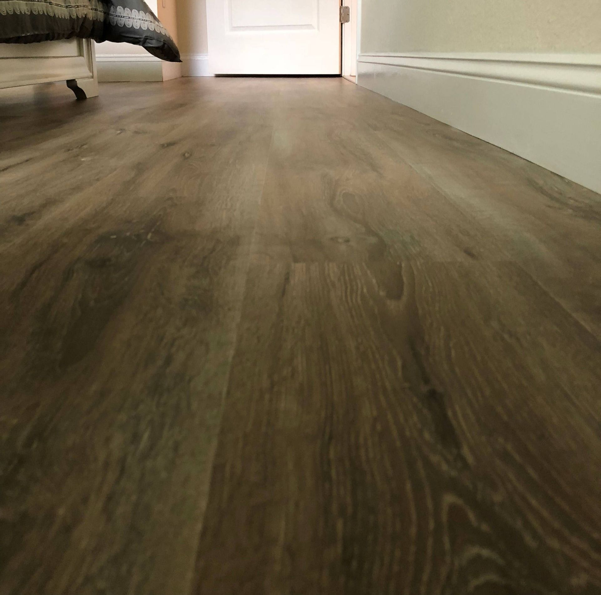 Low-angle view of a wood floor hallway leading to a white door, with a bed edge on the left