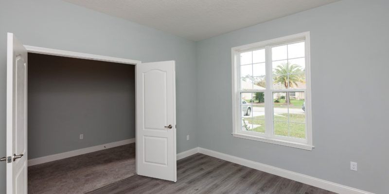 Empty light-gray bedroom with open double doors, dark carpet, and a window letting in daylight